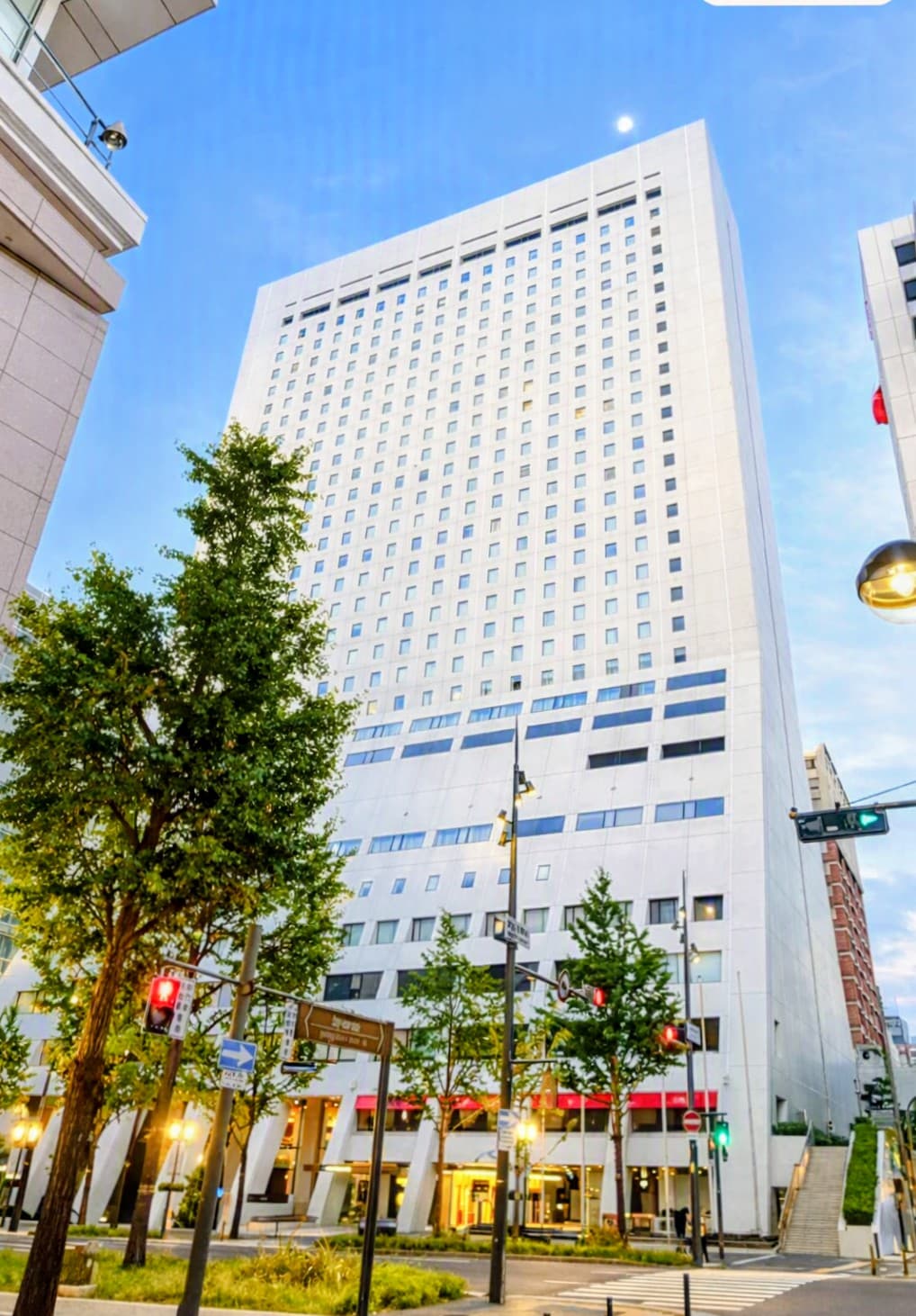 Tall white skyscraper with many windows rises above green trees on a bright city street.
